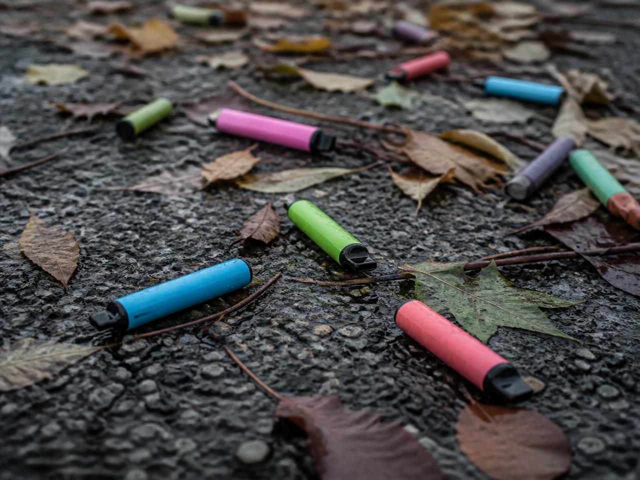 Weggeworfene bunte Einweg-E-Zigaretten zwischen Herbstblättern auf nassem Asphalt